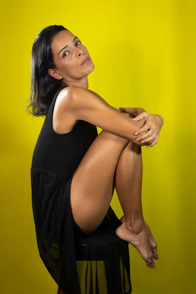 長い黒いドレスの美しいモデルは 彼女の足で木製のスツールに座って描かれた 黄色の背景に隔離された — ストック写真