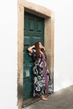Elleri başının arkasında, yeşil renkli bir kapıya dayanmış renkli elbiseli güzel bir kadın. Sessiz ve mutlu bir insan. Pelourinho, Brezilya.