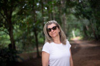 Ormanda güneş gözlüklü güzel bir kadın kameraya bakıyor. Ağaç yolu. Çevre.