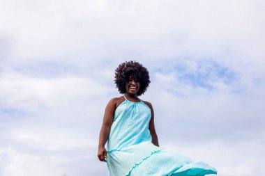 Arka planda mavi gökyüzüne karşı siyah saçlı, uzun mavi elbiseli güzel bir kadın portresi. Rio Vermelho Plajı, Salvador, Brezilya.