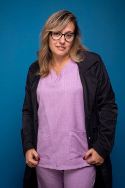 Siyah ceketli, eflatun üniformalı, gözlüklü, kameraya bakan kendine güvenen bir kadın doktor. Pozitif kişi. Mavi arkaplanda izole