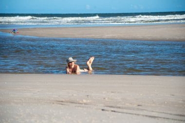 Bikinili ve şapkalı bir kadın denizin sularında ayaklarını kaldırıyor. Taquari, Guaibim plajı, Valenca, Bahia.