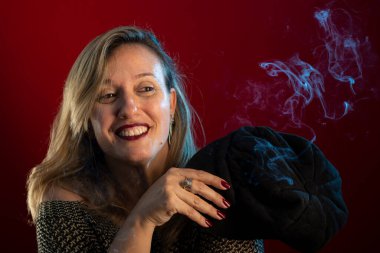 Çekici ve kendine güveni tam bir kadın, siyah bir bereliyi yüzünün yanında tutuyor. Tütsü dumanı her yerde. Kırmızı arkaplanda stüdyo portresi.