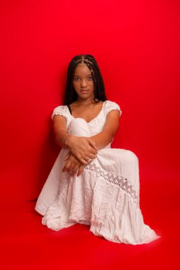 Güzel, genç, siyah bir kadın sakince oturuyor uzun örgülü beyaz elbiseler giyiyor. Kırmızı arkaplanda izole