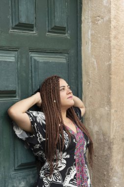 Elleri başının arkasında, yeşil renkli bir kapıya dayanmış renkli elbiseli güzel bir kadın. Sessiz ve mutlu bir insan. Pelourinho, Brezilya.