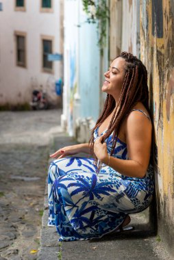 Saçları örgülü güzel bir kadın arka planda sokaklar olan bir duvara yaslanıyor. Pelourinho, Brezilya.