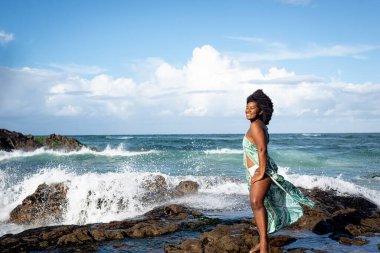 Siyah saçlı, gülümseyen, açık yeşil elbiseli, beyaz bikinili, kumsalın kayalarında duran güzel bir kadın. Mavi gökyüzü ve arka planda bulutlar. Rio Vermelho Plajı, Salvador, Brezilya.