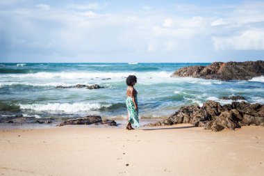 Güzel bir kadın kumsalda kumların üzerinde, sırtı kameraya dönük. Gökyüzü bulutları kayalar ve arka planda deniz. Rio Vermelho Plajı, Salvador, Brezilya.