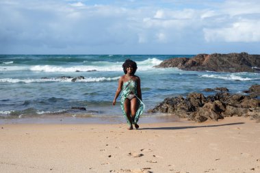 Kumsalda yürüyen, uzun elbiseli güzel bir kadın. Gökyüzü bulutları kayalar ve arka planda deniz. Rio Vermelho Plajı, Salvador, Brezilya.