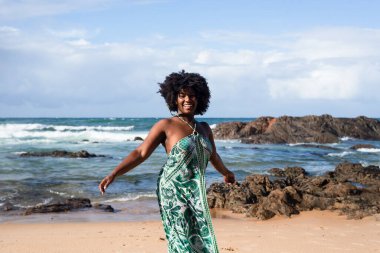 Kumsalda gülümseyen ve mutlu duran güzel siyah saçlı kadının portresi. Arka planda gökyüzü kayaları ve denizi bulutlandırır. Rio Vermelho, Salvador, Brezilya.