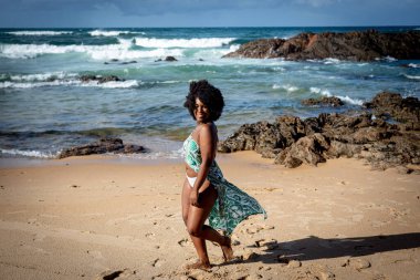 Kumsalın kumlarında oynayan siyah saçlı, mavi elbiseli güzel bir kadın. Mutlu ve kendinden emin biri. Arka planda gökyüzü kayalar ve deniz. Rio Vermelho Plajı, Salvador, Brezilya.