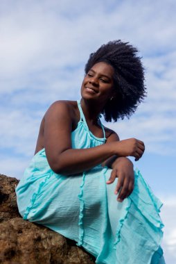 Plaj kayasında oturan siyah saçlı, mavi elbiseli güzel bir kadın. Kendinden emin ve pozitif bir kadın. Arka planda mavi gökyüzü ve bulutlar. Rio Vermelho Plajı, Salvador, Brezilya.