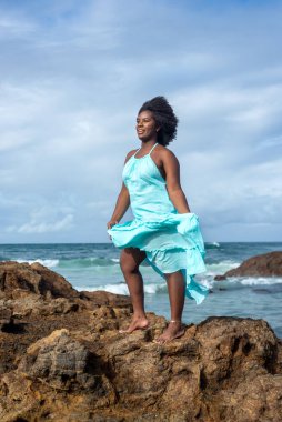 Kumsaldaki bir kayanın tepesinde siyah saçlı, mavi elbiseli bir kadın. Arka planda gökyüzü ve deniz. Rio Vermelho, Salvador, Brezilya.