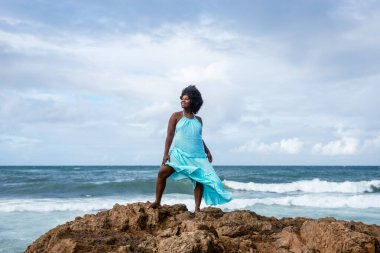 Siyah saçlı güzel bir kadının portresi kumsaldaki bir kayanın tepesinde mavi bir kıyafet giyiyor. Arka planda gökyüzü ve deniz. Rio Vermelho, Salvador, Brezilya.