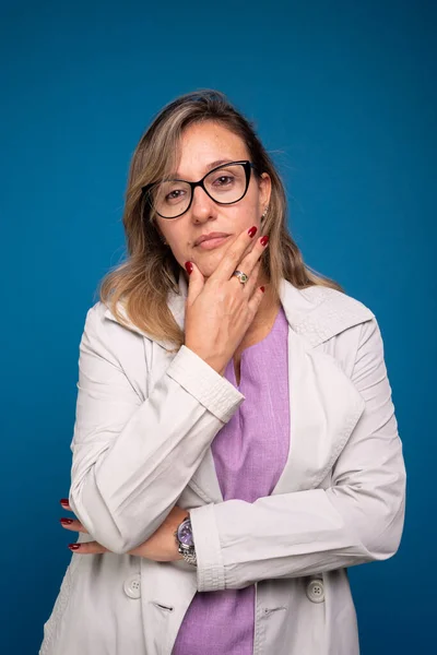 ベージュ色のコートに身を包んだ自信のある女性医師の肖像画で 眼鏡をかけ あごに手をつけている 肯定的な人 青い背景に隔離された — ストック写真
