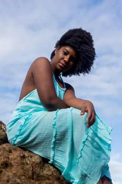 Beautiful woman with black power hair sitting on a beach rock wearing blue outfit. Confident and positive woman. In the background blue sky and clouds. Rio Vermelho Beach, Salvador, Brazil.
