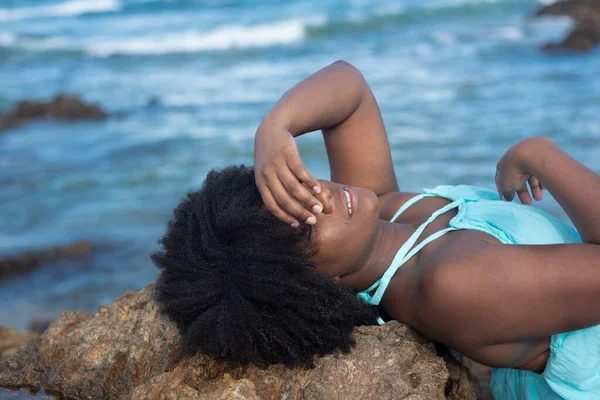 Beautiful woman with black power hair lying on the rocks of a beach In the background the waters of the sea. Rio Vermelho Beach, Salvador, Brazil.