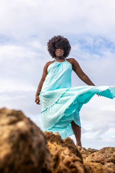 Happy beautiful woman with black power hair in light blue clothing on top of a rock. In the background sky and sea. Rio Vermelho, Salvador, Brazil.