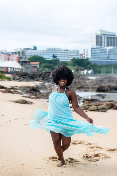 Beautiful woman with black power hair wearing long light blue outfit dancing on the beach sand. Happy and confident woman. Sky and clouds in the background. Rio Vermelho Beach, Salvador, Brazil.
