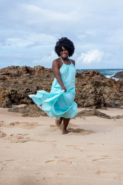 Beautiful woman with black power hair wearing long light blue outfit dancing on the beach sand. Happy and confident woman. Sky and clouds in the background. Rio Vermelho Beach, Salvador, Brazil.