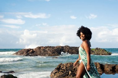 Siyah saçlı, gülümseyen, açık yeşil elbiseli, beyaz bikinili, kumsalın kayalarında duran güzel bir kadın. Mavi gökyüzü ve arka planda bulutlar. Rio Vermelho Plajı, Salvador, Brezilya.