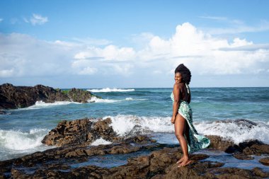 Siyah saçlı, gülümseyen, açık yeşil elbiseli, beyaz bikinili, kumsalın kayalarında duran güzel bir kadın. Mavi gökyüzü ve arka planda bulutlar. Rio Vermelho Plajı, Salvador, Brezilya.
