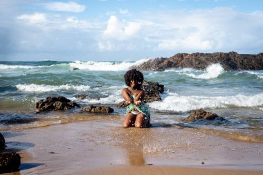 Siyah saçlı, kumsalda diz çöküp su atan güzel bir kadının portresi. Mavi gökyüzü kayalar ve arka planda bulutlar. Rio Vermelho Plajı, Salvador, Brezilya.