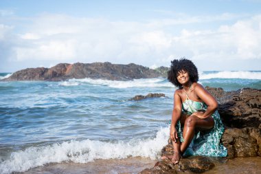 Plajda güneşlenen ve kameraya bakan uzun takım elbiseli kadın portresi. Arka planda gökyüzü bulutları ve kayalar. Rio Vermelho Plajı, Salvador, Brezilya.