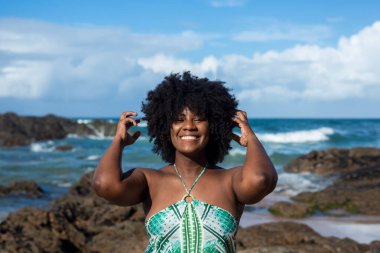 Siyah güç saçlarıyla oynayan güzel bir kadının portresi. Kendine güvenen ve mutlu gülümseyen. Arka planda gökyüzü kayaları ve denizi bulutlandırır. Salvador, Brezilya.