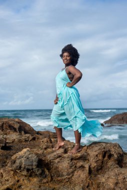 Siyah saçlı, kumsaldaki bir kayanın tepesinde mavi kıyafetli bir kadın portresi. Arka planda gökyüzü ve deniz. Rio Vermelho Plajı, Salvador, Brezilya.
