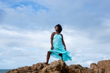 Siyah saçlı güzel bir kadının portresi kumsaldaki bir kayanın tepesinde mavi bir kıyafet giyiyor. Arka planda gökyüzü ve deniz. Rio Vermelho, Salvador, Brezilya.