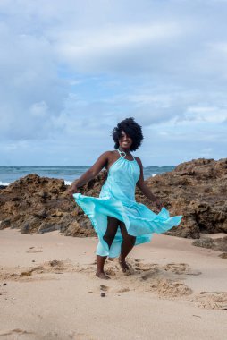 Siyah saçlı, açık mavi elbiseli, kumsalda dans eden güzel bir kadın. Mutlu ve kendinden emin bir kadın. Arka planda gökyüzü ve bulutlar. Rio Vermelho Plajı, Salvador, Brezilya.