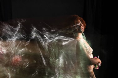 Kıvırcık kızıl saçlı, elinde bir buket çiçek tutan güzel bir kadının düşük hızlı çekimi. Açık renkli giysiler giyiyor. Siyah arkaplanda izole.