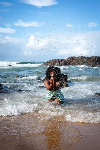 Portrait of beautiful woman with black power hair kneeling on beach sand throwing water up. Blue sky rocks and clouds in the background. Rio Vermelho Beach, Salvador, Brazil.