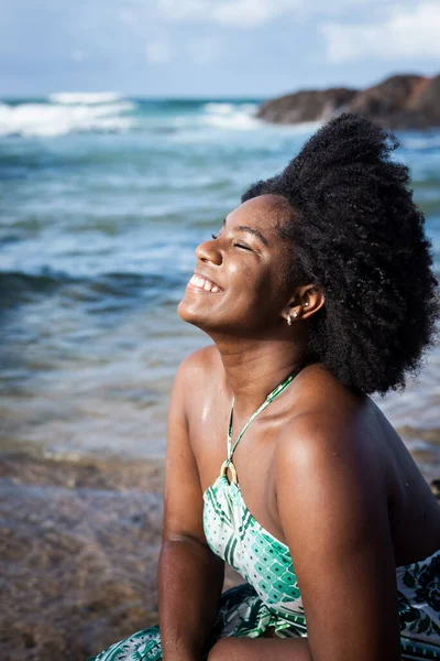 Beautiful woman with black power hair very happy and smiling looking at the camera. Sky clouds rocks and the sea in the background. Rio Vermelho Beach, Salvador, Brazil.