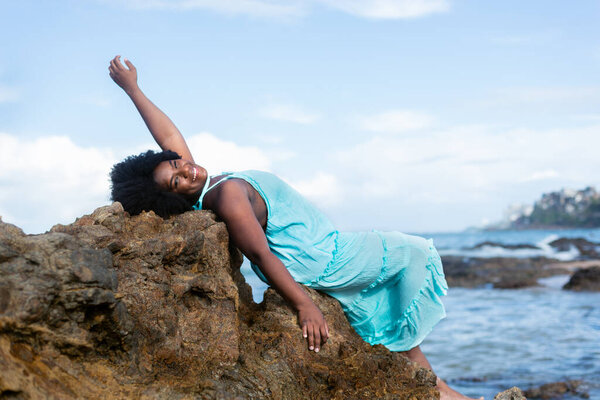 Portrait of a beautiful woman with black power hair lying on the rocks of a beach wearing light blue clothes. In the background blue sky clouds and the sea. Rio Vermelho Beach, Salvador, Brazil.
