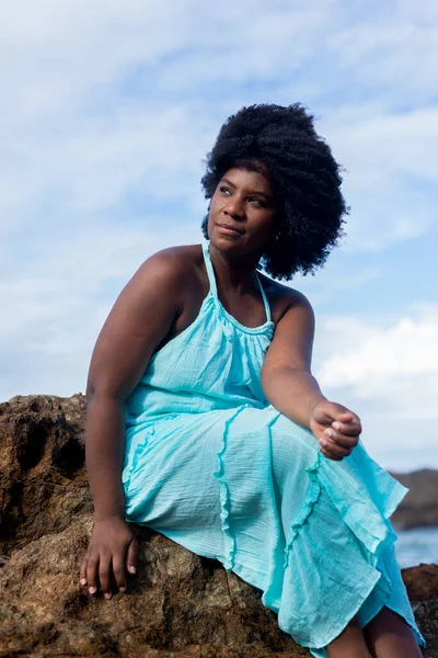 Beautiful woman with black power hair sitting on a beach rock wearing blue outfit. Confident and positive woman. In the background blue sky and clouds. Rio Vermelho Beach, Salvador, Brazil.