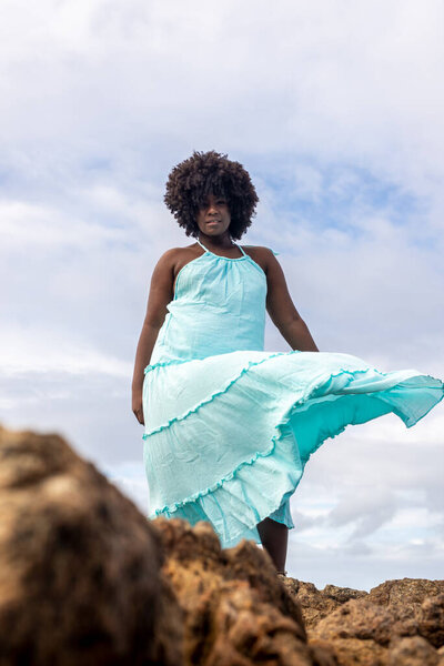 Happy beautiful woman with black power hair in light blue clothing on top of a rock. In the background sky and sea. Rio Vermelho, Salvador, Brazil.