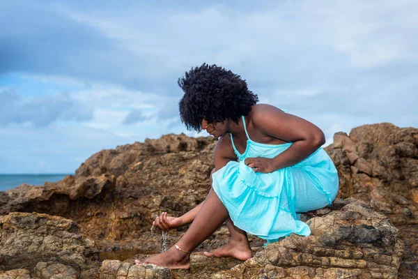A beautiful woman with black power hair in a long blue outfit sits on the beach rock touching her feet. Confident and happy person. Rio Vermelho Beach, Salvador, Brazil.