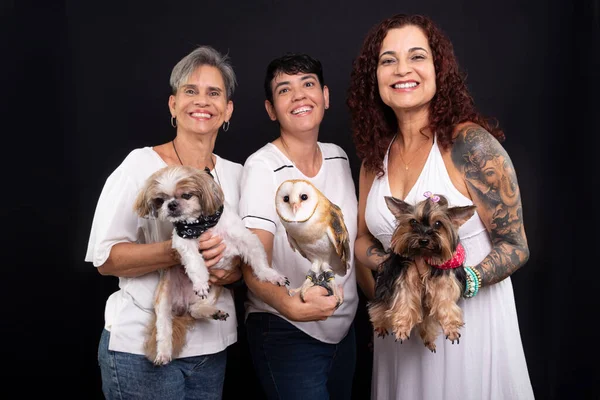 Three happy, confident lesbian women with their pets. An owl and Yorkshire and Shih Tzu dogs. Studio shot against black background.