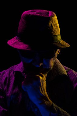 Yüzüne dokunan şapka ve güneş gözlüğü takan ciddi ve gizemli bir adamın portresi. Stüdyoda renkli ışıklar ve koyu arkaplan ile portre.