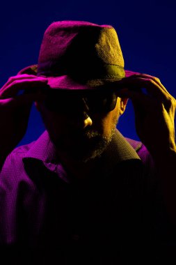 Şapka takan ve şapkasına güneş gözlüğü takan gizemli, ciddi bir adam. Stüdyoda renkli ışıklar ve koyu arkaplan ile portre.