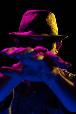 Şapka takıp güneş gözlüğü takan gizemli, ciddi sakallı adam elleriyle jestler yapıyor. Stüdyoda renkli ışıklar ve koyu arkaplan ile portre.