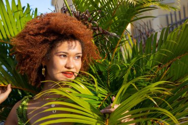 Bir bitkinin yaprakları arasında kızıl saçlı, öğleden sonra güneşi yüzüne vuran bir kadın. Kendinden emin ve mutlu biri..