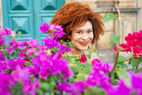 Portrait of a smiling red-haired woman among lilac and red flowers with green leaves. Confident and calm person.