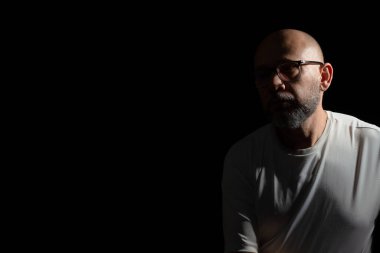 Siyah arka planda günlük kıyafetler giyen kel, sakallı bir adam..