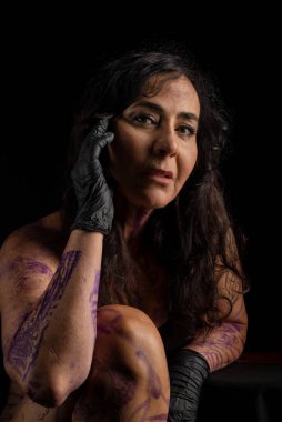 Dövmeli sanatçı, oturuyor, siyah arka planda boyalı ve dövmeli.