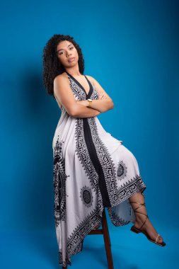 Tahta bir taburede oturmuş fotoğraf için poz veren kıvırcık saçlı güzel bir genç kadın. Mavi arkaplanda izole.