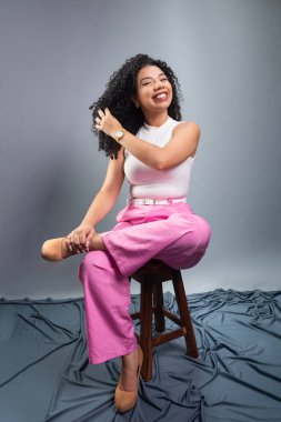 Kıvırcık saçlı, resmi giyinmiş güzel bir kadın tahta taburede oturmuş fotoğraf için poz veriyor. Gri arkaplanda izole.