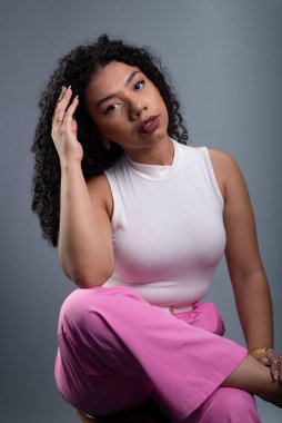 Güzel bir kadın oturuyor kıvırcık saçlarına dokunuyor ve gri arka planda kameraya bakıyor..